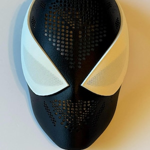May include: A black and white mask with a perforated design. The mask features white eye coverings and a black base with a pattern of small holes. The mask is likely for cosplay or costume use.