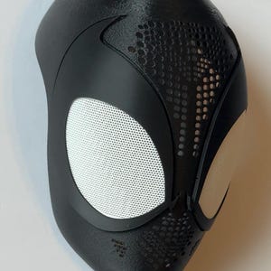 May include: Black superhero mask with white mesh eye coverings and a textured surface. The mask features a sleek design with perforated details near the eyes and mouth area, and is likely for cosplay or costume use.