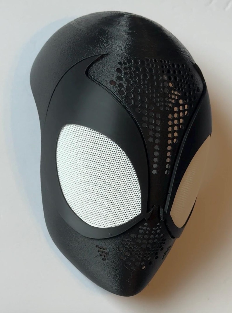 May include: Black superhero mask with white mesh eye coverings and a textured surface. The mask features a sleek design with perforated details near the eyes and mouth area, and is likely for cosplay or costume use.