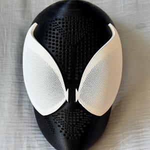 May include: A black and white mask with a textured surface. The mask features large white eye coverings with a mesh design. The black portion of the mask has a dotted pattern.