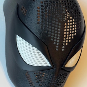 May include: Black superhero mask with white mesh eye coverings and a textured surface. The mask features a series of small, evenly spaced holes across the forehead and cheeks. The eye openings are outlined in black.
