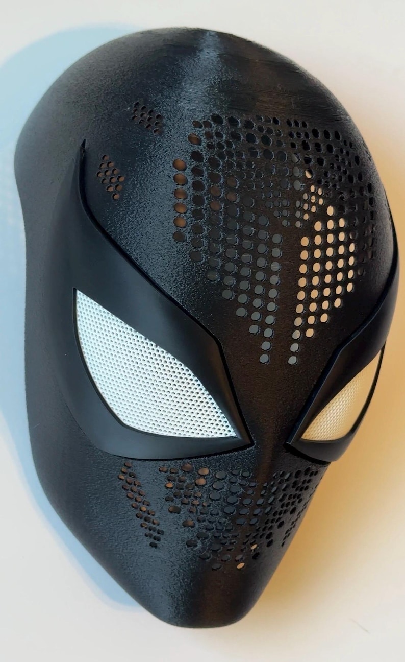 May include: Black superhero mask with white mesh eye coverings and a textured surface. The mask features a series of small, evenly spaced holes across the forehead and cheeks. The eye openings are outlined in black.