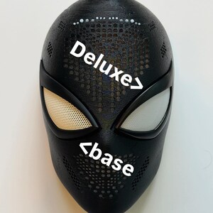 Op de afbeelding: Zwart masker met witte ooglenzen en de tekst "Deluxe" en "base". Het masker heeft een gestructureerd oppervlak met kleine gaatjes, waarschijnlijk een kostuumaccessoire.