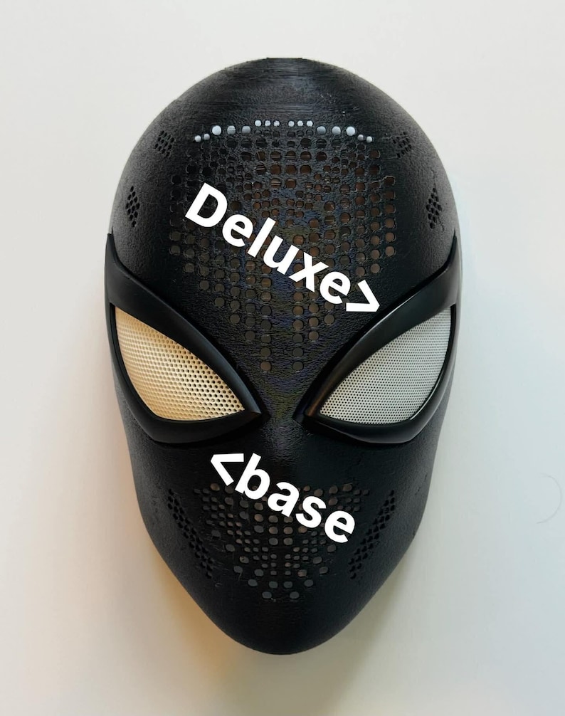Op de afbeelding: Zwart masker met witte ooglenzen en de tekst "Deluxe" en "base". Het masker heeft een gestructureerd oppervlak met kleine gaatjes, waarschijnlijk een kostuumaccessoire.