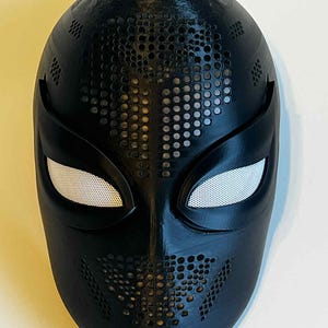 May include: A black mask with a textured surface and a series of small, evenly spaced holes. The eye openings are white with a mesh-like material. The mask has a sleek, modern design, suitable for cosplay or costume use.