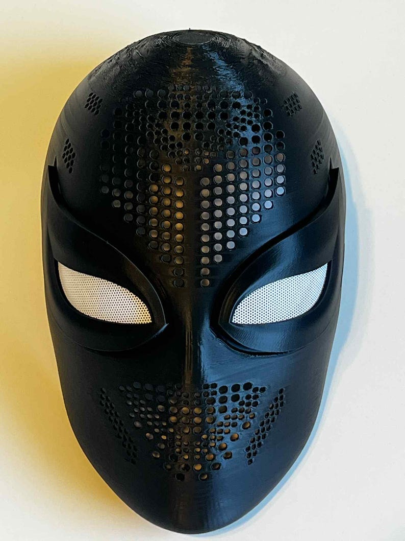 Puede incluir: Una m&aacute;scara negra con una superficie texturizada y una serie de peque&ntilde;os agujeros espaciados uniformemente. Las aberturas para los ojos son blancas con un material similar a una malla. La m&aacute;scara tiene un dise&ntilde;o elegante, adecuado para cosplay o disfraces.