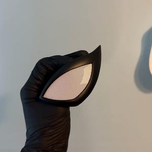 May include: A single eye lens piece for a mask, featuring a sharp, angular black frame surrounding a white mesh screen. The lens has a distinct almond shape with a pointed outer corner, designed with a smooth matte finish on the frame.
