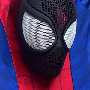 May include: A black full-face mask featuring a textured surface with a perforated pattern on the forehead and chin areas. The mask has two large, almond-shaped white mesh eye lenses framed by thick black borders, resting on a red and blue fabric.