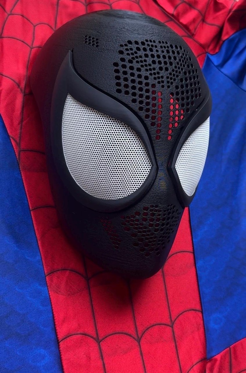 May include: A black full-face mask featuring a textured surface with a perforated pattern on the forehead and chin areas. The mask has two large, almond-shaped white mesh eye lenses framed by thick black borders, resting on a red and blue fabric.