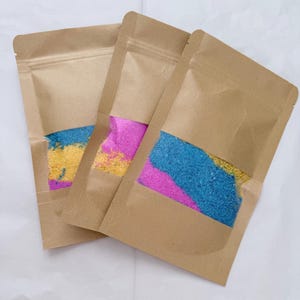 May include: Three brown paper pouches with clear windows, each containing colourful granular material. The colours include blue, yellow, and pink. The pouches are sealed with a zip closure.