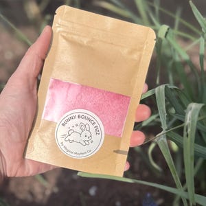 May include: A rectangular brown kraft paper pouch with a clear window revealing bright pink powder. A circular white label on the front features a black line drawing of a running rabbit and the text Bunny Bounce Fizz by Magical Mayhem Co.