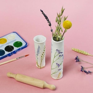 May include: Two white ceramic vases with floral designs, one holding dried flowers. A wooden rolling pin, watercolor paint set, and paintbrush are on a pink surface. The vases have lavender and green accents.