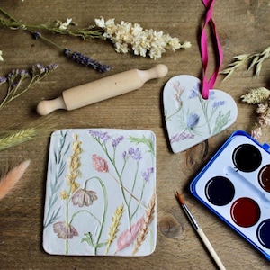 Può includere: Un mattarello di legno, un ornamento a forma di cuore e una piastrella rettangolare, tutti decorati con disegni floreali. Un set di acquerelli e un pennello sono anch'essi presenti. Gli oggetti sono disposti su una superficie di legno, con fiori secchi sparsi.