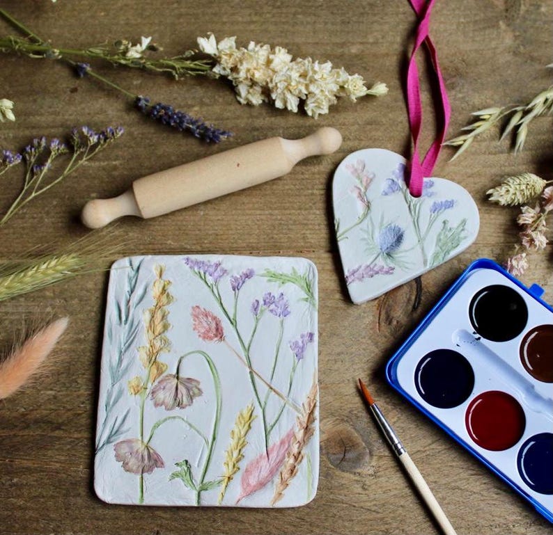 May include: A wooden rolling pin, a heart-shaped ornament, and a rectangular tile, all decorated with floral designs. A watercolor paint set and a paintbrush are also present. The items are arranged on a wooden surface, with dried flowers scattered around.
