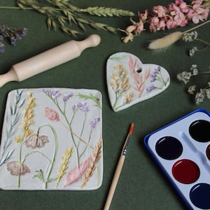 May include: An overhead shot features a wooden rolling pin, a heart-shaped and a rectangular ceramic piece, and a watercolor paint set. The ceramic pieces are decorated with floral designs in pastel colors. A paintbrush and dried flowers are also present.