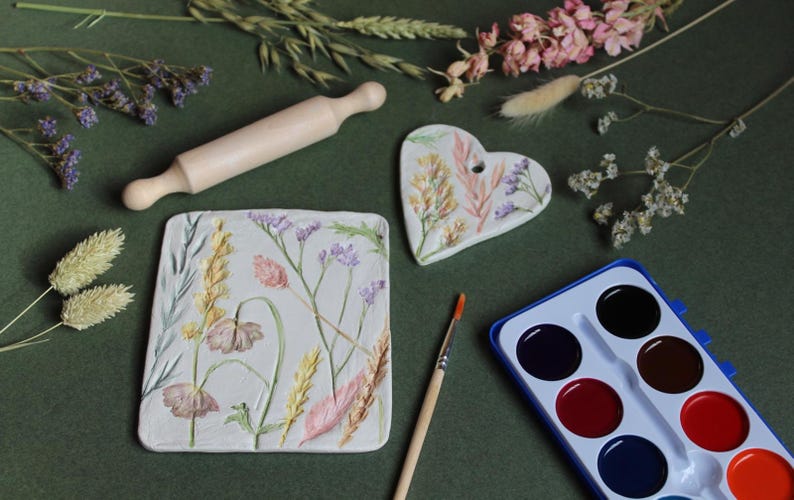 May include: An overhead shot features a wooden rolling pin, a heart-shaped and a rectangular ceramic piece, and a watercolor paint set. The ceramic pieces are decorated with floral designs in pastel colors. A paintbrush and dried flowers are also present.