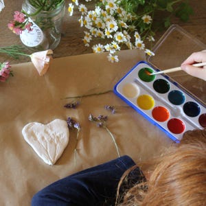 May include: A craft scene with a watercolor paint set, a heart-shaped clay piece, and various flowers. A person is using a paintbrush to apply green paint. The scene is set on a brown paper surface.