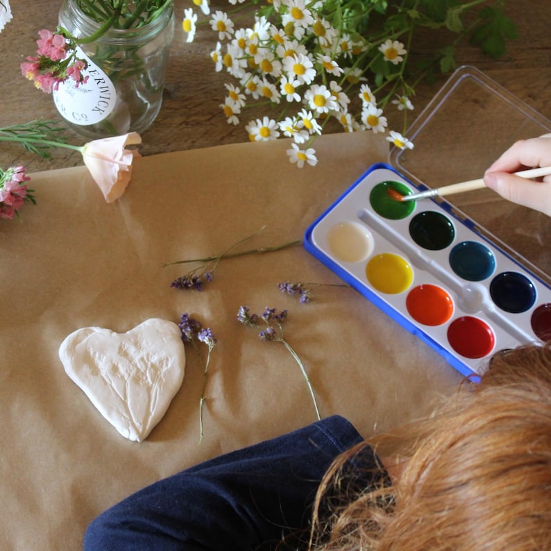May include: A craft scene with a watercolor paint set, a heart-shaped clay piece, and various flowers. A person is using a paintbrush to apply green paint. The scene is set on a brown paper surface.