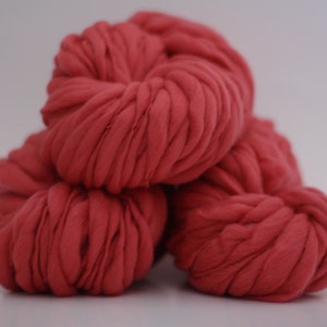 May include: Four balls of thick, coral-colored yarn are stacked against a white background. The yarn appears to be a chunky knit, with visible texture and a soft, inviting appearance. Ideal for crafting projects.