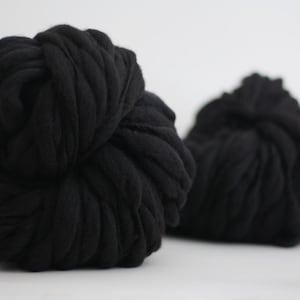 May include: Close-up of two large, black balls of thick yarn. The yarn is tightly wound, creating a textured surface. The background is plain white, highlighting the dark color and texture of the yarn.