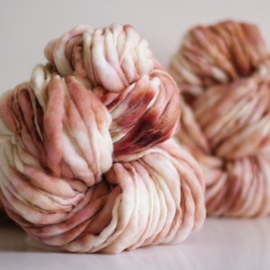 May include: Two skeins of hand-dyed yarn in shades of white, pink, and brown. The yarn is thick and fluffy, and it is perfect for knitting or crocheting.