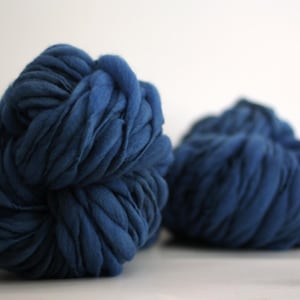 May include: Two large, rounded skeins of thick, chunky navy blue yarn. The yarn features a soft, roving-like texture with visible, voluminous strands coiled together to form dense, rounded balls of deep blue fiber.