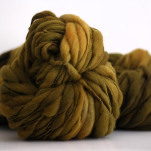 May include: A close-up of a bundle of chunky yarn in shades of olive green and brown. The yarn is soft and fluffy, and it has a slightly textured appearance.