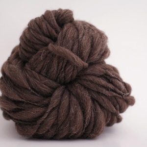 May include: A large ball of dark brown yarn, tightly wound and textured. The yarn appears thick and chunky, suitable for various crafting projects. The background is plain white, highlighting the yarn's color and form.