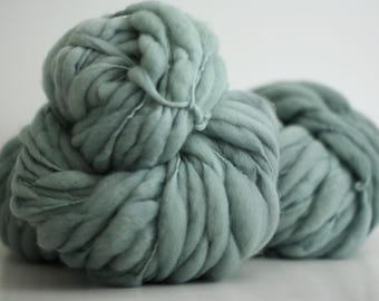 May include: A large skein of soft, chunky yarn in a muted sage green color. The yarn is loosely wound and has a fluffy, textured appearance.