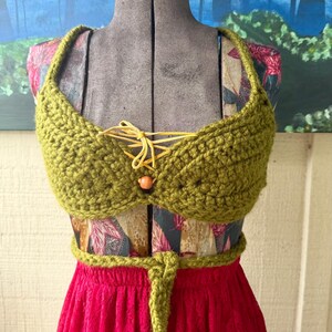 Op de afbeelding: Een handgemaakte olijfgroene gehaakte top met een veter aan de voorkant en een kleine houten kraal. De top wordt getoond op een mannequin over een stof met patroon en een felroze rok. De top heeft een bijpassende riem.