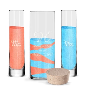 May include: A wedding sand ceremony set with three glass vases. Two vases are filled with coral and blue sand, labeled "Mr." and "Mrs." The center vase has blue and coral sand with the couple's names and wedding date.