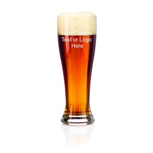 May include: A tall glass beer mug with a clear glass base and a frosted glass top. The mug is filled with a light brown beer and a white head of foam. The text "Your Custom Text or Logo Here" is printed on the side of the mug.