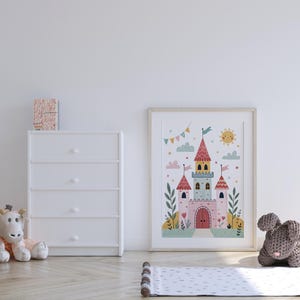 K&ouml;nnte beinhalten: Gerahmte Illustration eines skurrilen Schlosses mit rosa T&uuml;rmen, Flaggen und einer l&auml;chelnden Sonne. Das Kunstwerk wird neben einer wei&szlig;en Kommode, einer Stoffgiraffe und einem grauen gestrickten Hasen auf einem hellen Holzboden ausgestellt.