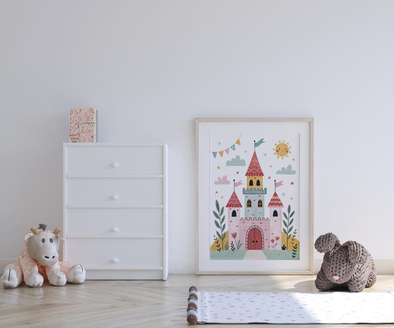 K&ouml;nnte beinhalten: Gerahmte Illustration eines skurrilen Schlosses mit rosa T&uuml;rmen, Flaggen und einer l&auml;chelnden Sonne. Das Kunstwerk wird neben einer wei&szlig;en Kommode, einer Stoffgiraffe und einem grauen gestrickten Hasen auf einem hellen Holzboden ausgestellt.