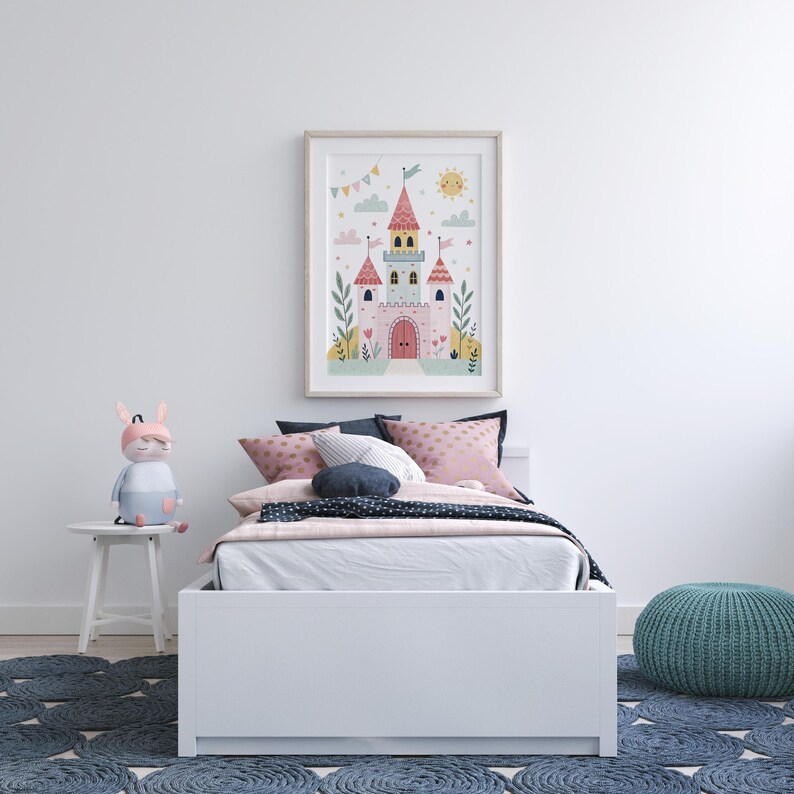 K&ouml;nnte beinhalten: Gerahmte Illustration eines rosa Schlosses mit roten T&uuml;rmen, Flaggen und einer Sonne. Das Kunstwerk h&auml;ngt &uuml;ber einem wei&szlig;en Bett mit rosa und blauen Kissen, einem blauen Teppich und einem Stoffhasenspielzeug auf einem kleinen wei&szlig;en Tisch.