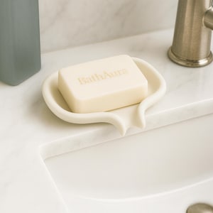 May include: A cream-coloured soap dish holds a rectangular bar of soap, with the text "Bath Aura" printed on it. The dish has a curved, organic shape and sits on a white countertop. A tap is visible in the background.