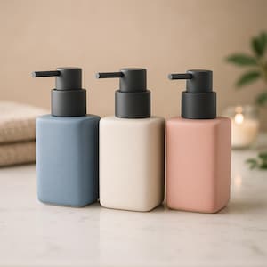 Puede incluir: Tres dispensadores de jabón de cerámica en azul, crema y rosa. Cada uno tiene una bomba gris oscuro y un cuerpo rectangular. Los dispensadores están dispuestos sobre una superficie blanca, lo que sugiere un baño o una cocina. El diseño es minimalista y moderno.