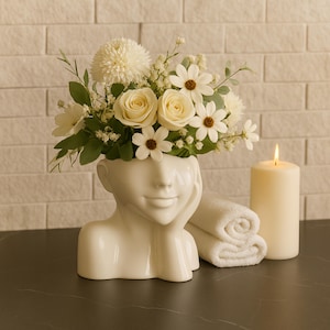 Può includere: Un vaso bianco a forma di testa pieno di fiori bianchi, tra cui rose e margherite. Una candela accesa e asciugamani bianchi arrotolati sono accanto al vaso. Lo sfondo è un muro di mattoni bianchi.