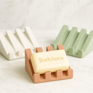May include: A close-up shot of three soap dishes in different colors: white, green, and peach. A bar of soap with the text "Bath Aura" rests in the peach-colored dish. The dishes have a slotted design.