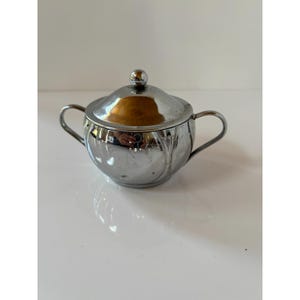 May include: A polished chrome sugar bowl with a rounded body, two curved side handles, and a matching lid featuring a small spherical knob. The metallic surface has a reflective finish that shows subtle vertical embossed lines along the sides.
