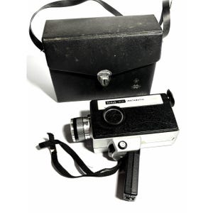 May include: A black and silver Kodak M30 Instamatic movie camera with a black textured grip, a lens, and a folding handle, accompanied by a matching black rectangular carrying case with a shoulder strap and a metal latch closure.
