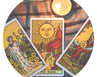 May include: A circular image featuring tarot cards and a lit candle. The central card displays a radiant sun with the text "THE SUN." Other cards show figures and symbols. The scene is set on a textured, neutral-toned surface.