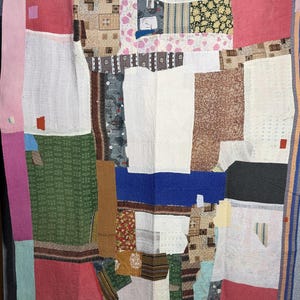 May include: A large rectangular quilt constructed from various fabric patches in diverse colours including red, green, blue, white, and brown. The textile features intricate running stitch patterns throughout, with a multi-coloured striped border.