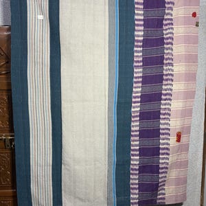 Puede incluir: Una colcha textil rectangular con paneles verticales de anchos variables. El diseño incluye rayas en tonos verde azulado, crema, azul oscuro y morado, con una superficie texturizada creada por finas y densas puntadas de costura.