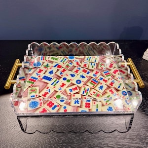 May include: A clear acrylic tray with a scalloped edge and gold handles, filled with ivory-coloured Mahjong tiles featuring red, blue, and green symbols. The tiles are arranged on a pink surface.