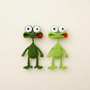 May include: Two handmade crochet frog toys. One is dark green, the other is light green. Both have large white eyes with black pupils, open mouths with red tongues, and small arms and legs. The frogs are positioned on a light beige background.