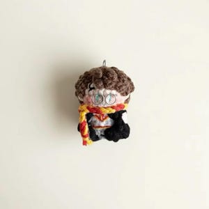 May include: A handmade crochet doll featuring a character with brown hair, round glasses, and a red and yellow scarf. The doll is wearing a black robe and a white shirt. A silver loop is attached to the top for hanging.