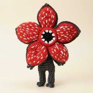 May include: A handmade crochet Demogorgon figure. The figure features a large red flower-like head with white accents and a black center. The body and legs are dark gray, with small feet. The figure is a unique, handcrafted item.