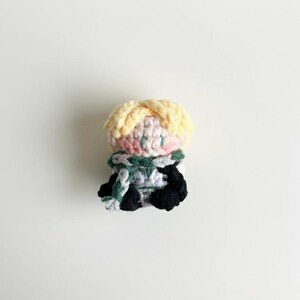 May include: A handmade crochet doll with a light yellow head, pink cheeks, and green eyes. The doll wears a green, white, and black scarf and a black outfit. The doll is sitting on a white surface.