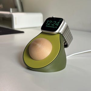 Könnte beinhalten: Eine Ladestation in Form einer Avocado mit einer hellgrünen, texturierten Mitte und einer glatten, hellen Holzkugel, die den Kern darstellt. Eine silberne Smartwatch mit einem Metallgliederarmband liegt oben auf dem Avocado-Ständer.
